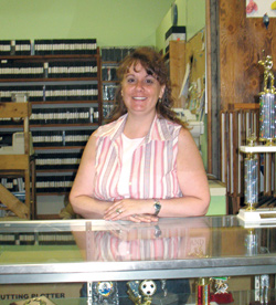 Photo by Amy Veneziano	Stephanie Bradley stands behind a new trophy case installed recently at II Sisters Video Store. The Bradley’s bought the store and added the trophy and sign-making business in April. 