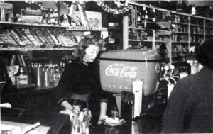 From All Roads Lead to Tonasket: "This interior picture was given by Lloyd Curtis. It was reproduced from a slide taken about 1956 when both the drug stores in town had soda fountains. Bev Haney is shown waiting on and unknown customer. Business was not too good for the fountains, so this one was taken out about 1958. The other one was also removed about the same time and there hasn't been one since."