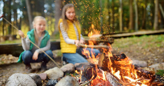 Young sisters roasting hotdogs on sticks at campfire. Adobe Free Stock.