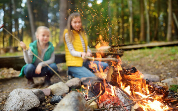 Young sisters roasting hotdogs on sticks at campfire. Adobe Free Stock.